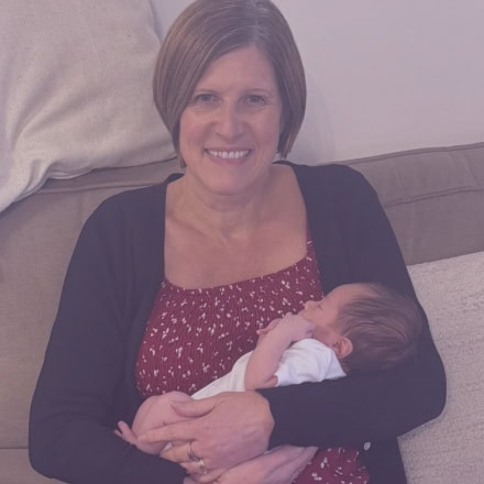Maria the Doula sitting on a sofa smiling whilst soothing and cuddling a newborn during a postnatal doula session.
